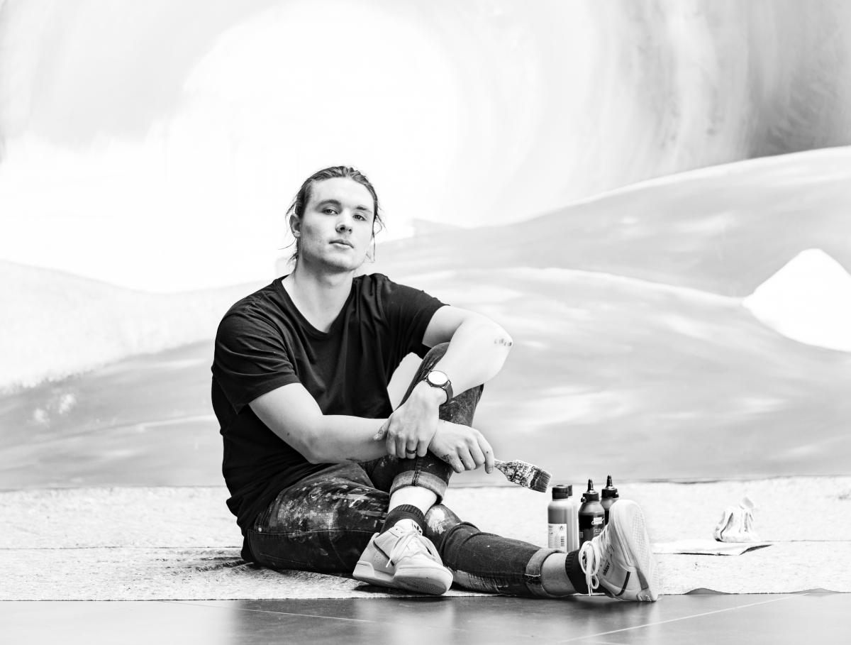 Der Künstler Simon Kohler mit Pinsel und Farben vor einem Gemälde sitzend (Schwarz-Weiß-Foto)